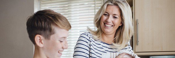 Smiling woman washes up in the kitchen of her Guernsey home, helped by her young son.