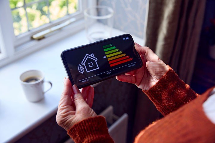 Senior woman holding a smart metre showing her home’s energy rating while trying to keep warm by a radiator. 