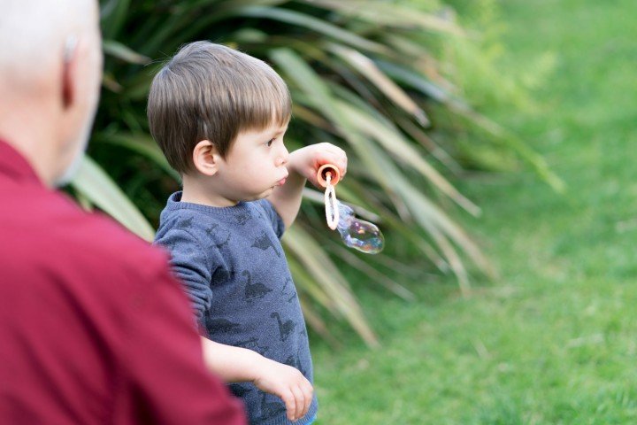 A young child with their grandparent blowing bubbles outdoors. 