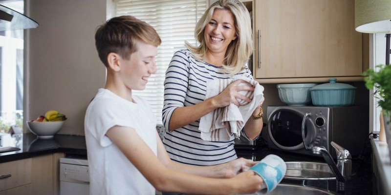 Smiling woman washes up in the kitchen of her Guernsey home, helped by her young son.